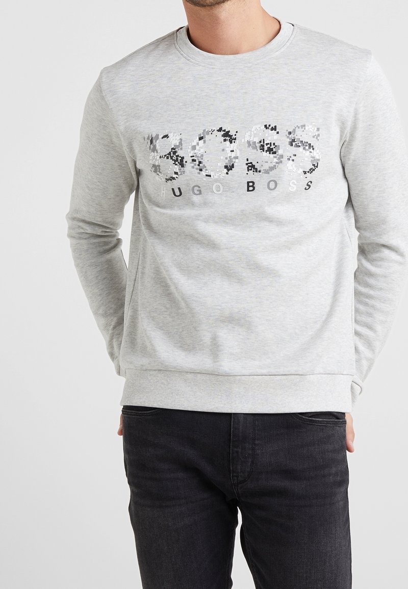 Man i ljusgrå långärmad Hugo Boss sweatshirt med pixelerad logotyp och svarta jeans, med händerna gömda bakom ryggen.
