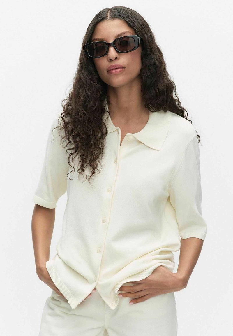 Kvinde med langt, krøllet hår iført sorte solbriller og en cremefarvet kortærmet skjorte med knapper samt matchende bukser, hænderne i lommerne.
