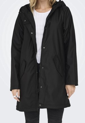 Femme portant un imperméable à capuche noir avec fermeture éclair et boutons-pression, par-dessus un haut blanc texturé et un pantalon noir avec détail côtelé.