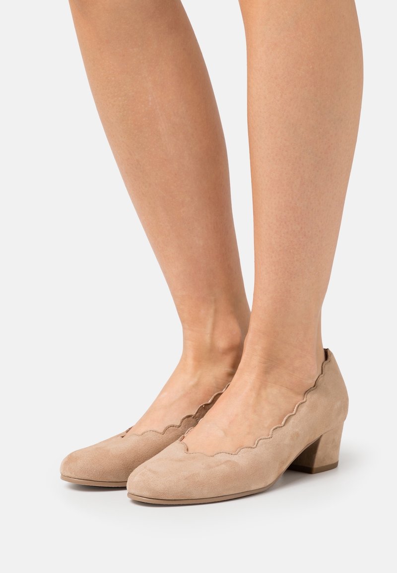 Chaussures beiges en daim à petit talon avec bords festonnés portées pieds nus sur un fond uni.
