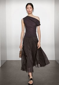 Donna che cammina sulla passerella indossando un top nero a spalla unica, una gonna nera fluida, sandali neri e teneva una piccola borsa di paglia marrone.