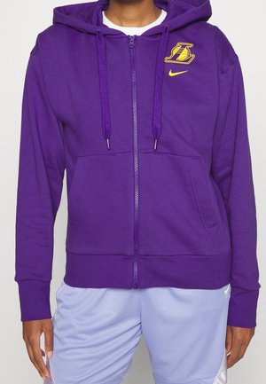 Osoba ubrana w fioletową bluzę na zamek z logo Los Angeles Lakers i Nike, w połączeniu z jasnofioletowymi spodenek sportowych.