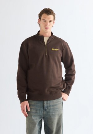 Jeune homme portant un sweat-shirt marron foncé à fermeture éclair quart avec un logo jaune "Wrangler" et un jean bleu clair sur un fond blanc uni.
