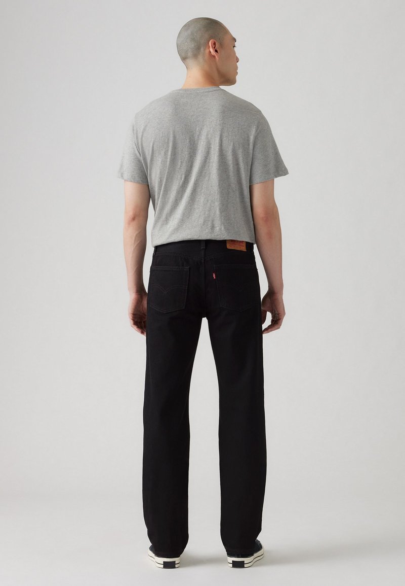 Grå kortärmad t-shirt, svarta raka jeans och svarta sneakers med vita sålar; modellen visas bakifrån.