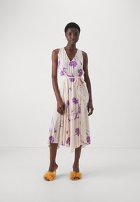 Abito midi floreale crema con stampa floreale viola, scollatura a V, vita arricciata con fiocco e gonna fluente. Abbinato a sandali aperti di pelliccia arancioni.