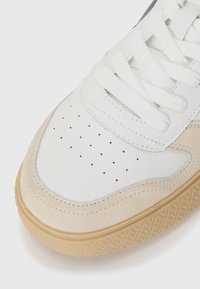 Hvit lær sneaker med beige semsket detaljer, perforert tåboks og hvite snøringer. Gummisålen har et teksturert mønster for bedre grep.
