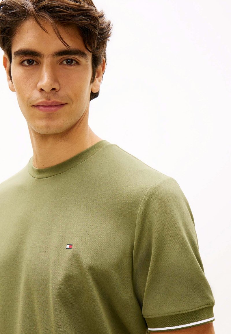Jeune homme aux cheveux courts et bruns portant un t-shirt vert olive à col rond avec un petit logo rectangulaire sur la poitrine.