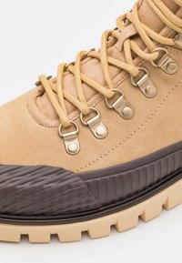 Bottes de randonnée en daim couleur fauve avec des lacets jaunes, des œillets en métal brossé, et une semelle en caoutchouc foncé texturée avec des crampons prononcés.