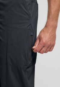 Donkergrijze broek met een ritszak en een gladde textuur. Een hand is te zien die de rits opent, wat de functionaliteit en het ontwerp benadrukt.