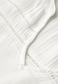White textured fabric with vertical pleats and delicate cutout detailing. Smooth surface and soft appearance.