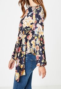 Blouse à fleurs avec un fond bleu marine foncé, manches longues bouffantes et un détail noué à la taille, confectionnée dans un tissu léger.