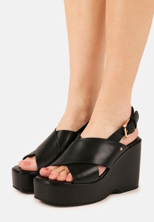 Pieds portant des sandales compensées en cuir noir avec de larges brides croisées et une bride à la cheville avec une boucle dorée, sur un fond clair uni.