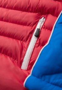 Close-up van een rood-blauw doorgestikt jack met een zichtbare witte ritssluiting en een zwart ritssluitlipje.