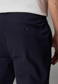 Pantalon en coton bleu marine avec une texture lisse, présentant une poche arrière et une fermeture à bouton, conçu pour un ajustement sur mesure.