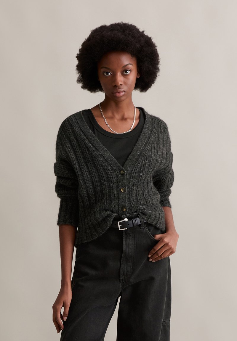 Donkergroene geribde cardigan met een V-hals, knopen aan de voorkant en oversized mouwen. Gemaakt met een zwarte top en hooggepaste broek.