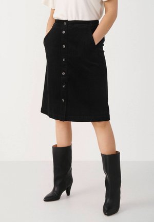 Femme portant une jupe en velours côtelé noire boutonnée devant avec des poches et des bottes noires à talons pointues mi-mollet, debout les mains dans les poches.
