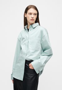 Mujer con cabello castaño lacio, lleva una camisa suelta de color verde menta con puños largos y pantalones de cuero negro, de pie contra un fondo blanco.