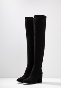 Paire de bottes en daim noir montant au-dessus du genou, avec talons blocs et bouts pointus, se tenant debout sur une surface blanche contre un fond uni.
