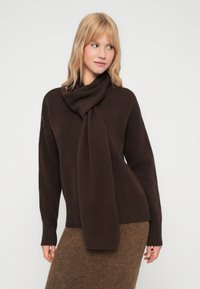 Mulher loira a usar um suéter castanho escuro e um cachecol a condizer, combinado com uma saia texturizada castanha, de pé contra um fundo claro e plain.