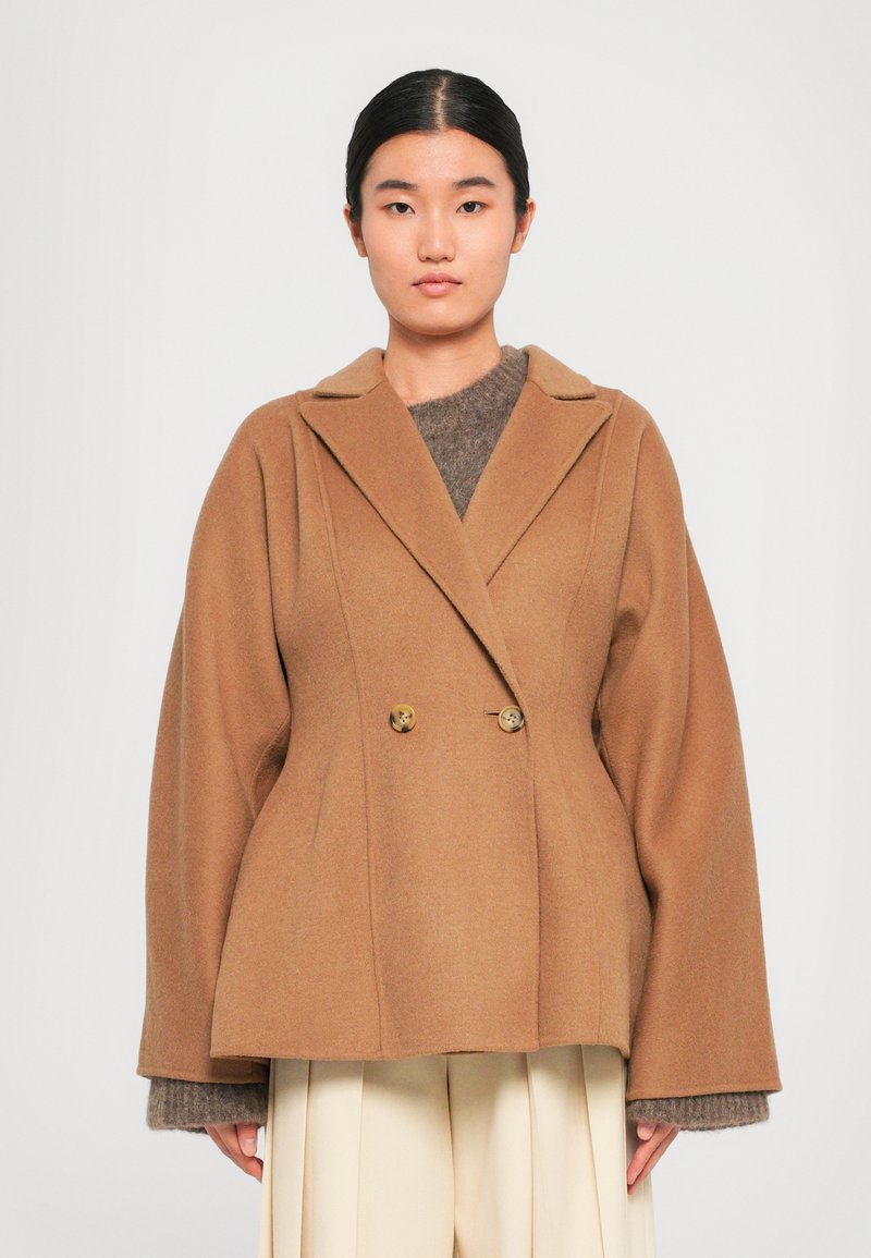 Manteau croisé marron en tissu doux, avec un col à revers et des manches larges. Deux boutons dorés ornent le devant.