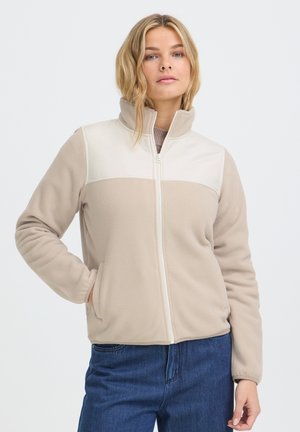 Femme portant une veste polaire beige et blanche à fermeture éclair, les mains dans les poches, debout devant un fond uni clair.