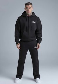 Ensemble sweat à capuche et pantalon de survêtement noirs en tissu doux. Le sweat à capuche comporte une fermeture éclair frontale et une poche, avec un logo discret. Des baskets blanches complètent le look.