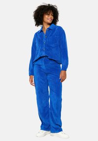 Modra kordna srajca z gumbastim zapiranjem in dvema žepoma, v kombinaciji z usklajenimi hlačami. Obe delu imata mehko teksturo in sproščen kroj.