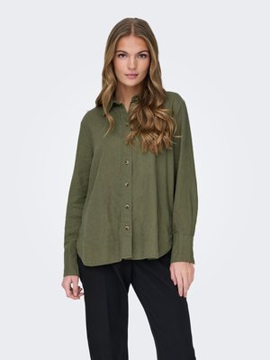 Femme aux longs cheveux ondulés portant une chemise boutonnée vert olive et un pantalon noir, debout devant un fond clair uni.