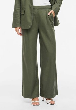 Kvinde iført olivengrønne bukser med vide ben og matchende blazer, hvid top og hvide åbne hælede sandaler, stående med den ene hånd i lommen.