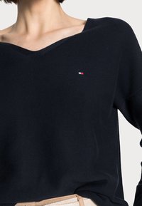 Pull en tricot bleu marine avec un col en V, arborant un petit logo rouge, blanc et bleu sur la poitrine et une coupe ample et décontractée.
