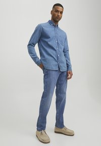 Camisa de mezclilla azul claro con mangas largas y cuello abotonado, combinada con vaqueros azul medio y zapatos beige tipo slip-on.