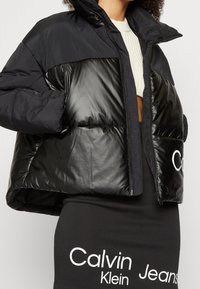 Svart kort puffjacka med glänsande paneler, hög krage, dragkedjestängning och en Calvin Klein-logotyp på sidan. Mjuk, quiltad textur.