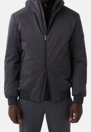 Mężczyzna w ciemnoszarej kurtce zapinanej na zamek z prążkowanymi mankietami i obrębem, zapinanymi na zamek bocznymi kieszeniami oraz kapturem, w połączeniu z szarymi spodniami.