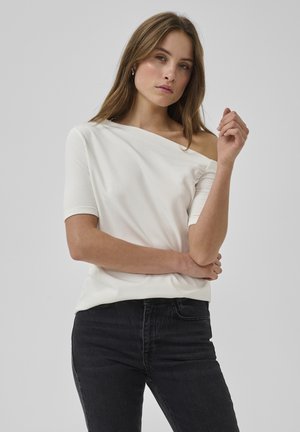 Valge õlapaeltega pluusi ja mustade teksadega naine heleda pruunide juustega, seisab ühe käega rüpes ja teine käsi on tõstetud.