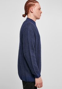 Yksi iso sininen neulepaita, jossa on ribbitekstuuri ja korkea kaulus. Sissä on laskeutuvat olkapäät ja pitkät hihat.