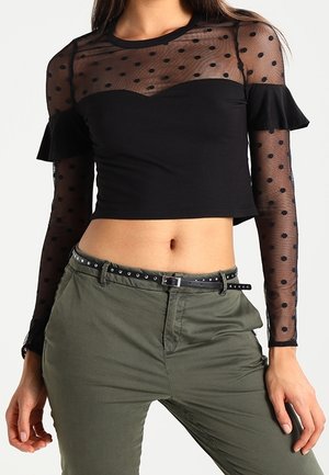 Femme portant un crop top noir transparent à pois, manches longues avec des volants, et un pantalon vert olive avec une ceinture noire cloutée.