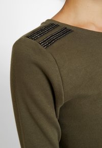 Suéter verde oliva con un diseño ajustado, textura acanalada y tres filas de adornos de perlas negras en los hombros.