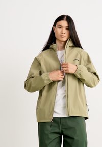 Jachetă de culoare verde oliv deschis cu fermoar în față, manșete elastice și detalii ale logo-ului. Asortată cu un tricou alb și pantaloni verzi închiși.
