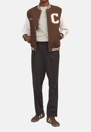 Bruine wollen varsityjacket met witte mouwen, voorzien van een grote letter "C," gecombineerd met een grijs shirt, zwarte broek en bruine sneakers.