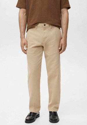 Mann trägt beige gerade geschnittene Hosen, schwarze Lederschuhe und ein braunes, strukturiertes Kurzarmhemd vor einfachem Hintergrund.