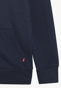 Sudadera azul marina hecha de una suave tela texturizada. Cuenta con un bolsillo frontal y puños de canalé, y tiene una pequeña etiqueta de logo rojo en la costura inferior.