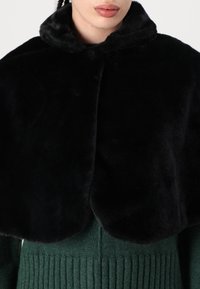 Svart kappa av konstpäls med en rundad krage och mjuk textur. Bärs över en grön ribbad tröja, vilket framhäver kontrasterande färger.
