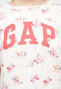 Chemise blanche avec de grandes lettres rouges "GAP" sur un imprimé floral multicolore, visible de la poitrine jusqu'au cou.