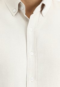 Camicia button-up beige chiaro con tessuto testurizzato, caratterizzata da un colletto a punta e cinque bottoni bianchi lungo la patta.