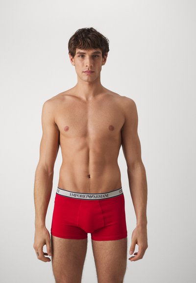 Röda kalsonger i boxermodell med en svart och vit märkesresår. Tyget verkar slätt, med en åtsittande form och minimala sömnadsdetaljer.