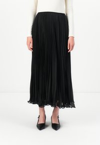 Mulher vestindo uma saia longa preta plissada, uma blusa branca de manga longa e sapatos de salto agulha pretos, de pé contra um fundo branco.
