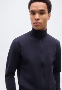 Νέος άνδρας που φοράει μια ναυτική μπλε ζακέτα με φερμουάρ και ψηλό γιακά, με το λογότυπο "BOSS" στο αριστερό στήθος, σε φόντο απλό και φωτεινό.