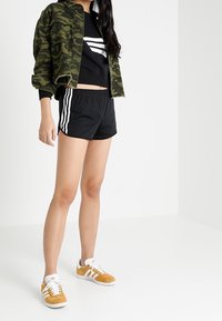 Camo jacket, black cropped sweatshirt with white stripes, black shorts, and yellow suede sneakers with white accents.