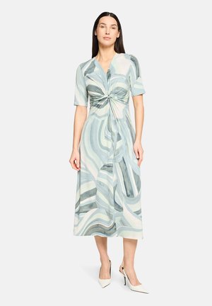 Maxikleid mit verdrehtem Vorderteil, kurzen Ärmeln und fließendem Design. Farben umfassen Hellblau, Grün und Creme mit Wellenmustern.