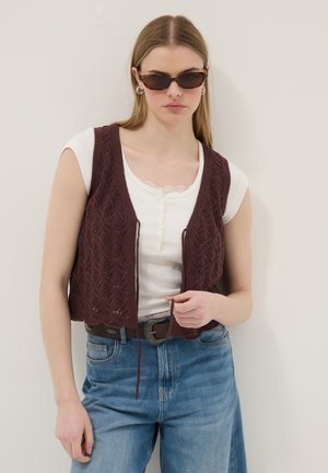 Junge Frau trägt Schildpatt-Sonnenbrille, weißes Kurzarmtop, braune Strickweste, blaue Jeans und Gürtel, steht vor einer hellen Wand.